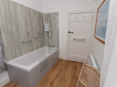 A bathroom with a bathtub shower combo wooden floor a white door a wicker chair and a wall picture at Armstrong Cottages No2 in Bamburgh