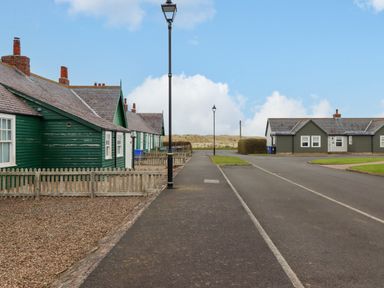 A street with green and gray houses along a paved road and a sidewalk at Armstrong Cottages No2 Bamburgh