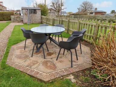 An outdoor patio with a round glass table and six black chairs surrounded by a wooden fence and garden sheds at Armstrong Cottages No2 Bamburgh