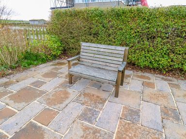 A wooden bench on a tiled patio with green shrubs behind at Armstrong Cottages No2 in Bamburgh
