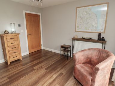A room with wooden flooring a wooden door a chest of drawers a small table a console table with decorative items a map on the wall and a velvet armchair at Assisi Apartment in Alnmouth