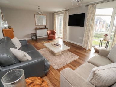 A living room with two sofas a coffee table a wall-mounted TV and large windows at Assisi Apartment in Alnmouth