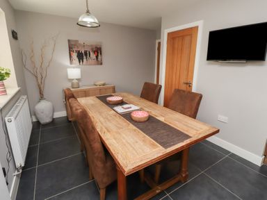 A dining room with a wooden table and brown chairs a sideboard with a lamp and artwork a wall-mounted television at Assisi Apartment in Alnmouth