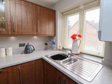 A kitchen corner with wooden cabinets a kettle canisters a double sink and a window with blinds and flowers at Assisi Apartment in Alnmouth