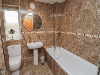 A bathroom with a toilet sink round mirror and bathtub with tiled walls at Assisi Apartment in Alnmouth