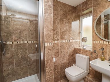 A bathroom with a shower, toilet, sink, round mirror, and a window at Assisi Apartment in Alnmouth