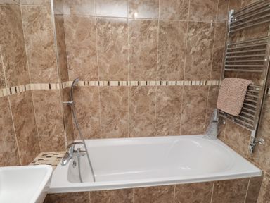 A bathroom with a white bathtub brown tiled walls a metal towel rack with a beige towel and a handheld shower head at Assisi Apartment in Alnmouth