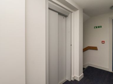 A hallway with a closed elevator door a fire exit sign a fire alarm and stairs at Assisi Apartment in Alnmouth