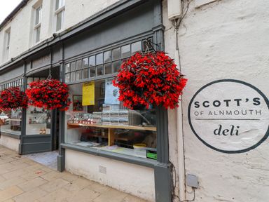 The exterior of Scotts of Alnmouth deli with hanging red flower baskets and a display window
