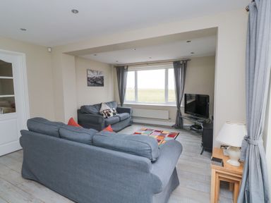 A living room with two gray sofas a television on a stand a window with curtains and a small table with a lamp at Beach View in Seahouses