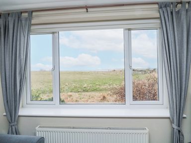 A window with gray curtains framing a view of a grassy field and blue sky at Beach View in Seahouses
