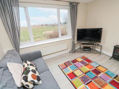 A living room with a gray sofa and patterned cushions a colorful rug a TV on a metal stand and a large window with a view at Beach View in Seahouses