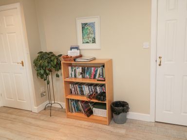 A room corner with a wooden bookshelf filled with books and DVDs a plant on a metal stand a wastebasket and two white doors at Beach View in Seahouses