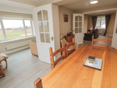A dining area with wooden table and chairs leading to a sitting room with sofas and a chair by large windows at Beach View in Seahouses