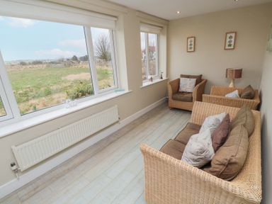 A sitting area with wicker furniture and cushions near large windows overlooking a grassy field at Beach View in Seahouses