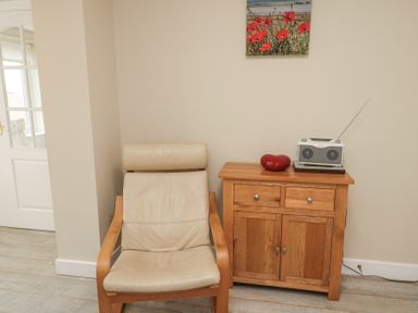 A beige armchair next to a wooden cabinet with a radio and a red decorative object on top and a poppy flower picture on the wall at Beach View in Seahouses