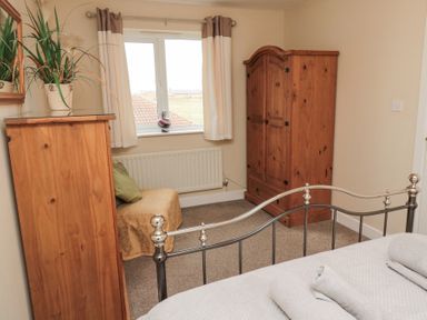 A bedroom with a metal bed frame, wooden wardrobe, chest of drawers, window with curtains, and a chair covered with a cloth at Beach View in Seahouses