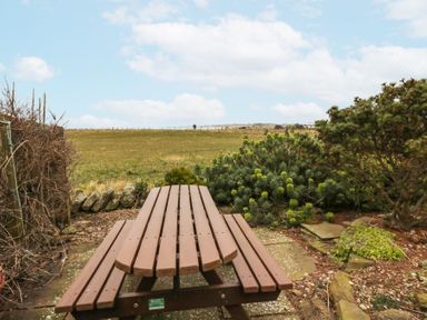A wooden picnic table with benches in a garden overlooking a grassy field and cloudy sky at Beach View in Seahouses