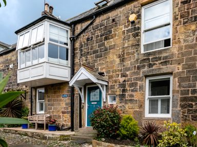 A stone house exterior with a blue front door and white framed windows with plants in front at Begonia House Alnmouth