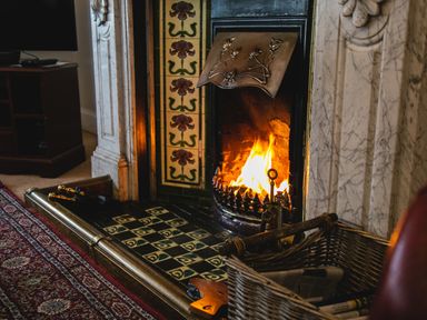 A lit fireplace with ornate tiles and a wicker basket nearby at Begonia House in Alnmouth
