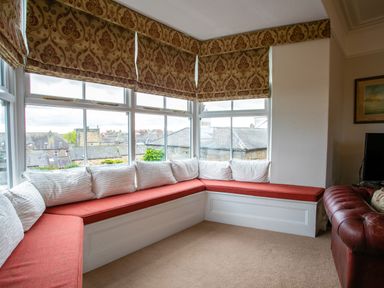 A corner window seating area with red cushions and white pillows next to a brown leather sofa at Begonia House in Alnmouth