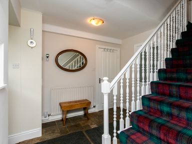 A hallway with a staircase covered in tartan carpet a wooden bench an oval mirror and a white door at Begonia House in Alnmouth