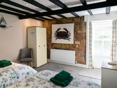 A bedroom with two beds with green towels and floral bedding a grey chair a cream wardrobe a crab painting on a stone wall and a window with striped curtains at Begonia House Alnmouth