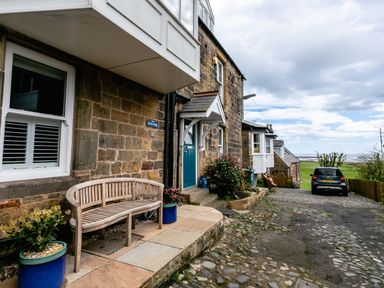 A stone house entrance with a wooden bench and potted plants on a cobblestone driveway with a parked car and view of the sea at Begonia House in Alnmouth