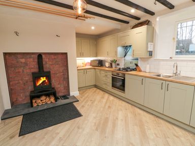 A kitchen with wooden cabinets a built in oven a fireplace with a fire burning and a window at Begonia House in Alnmouth