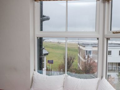 A window with raindrops and white cushions on the sill overlooking a grassy area and buildings at Begonia House in Alnmouth