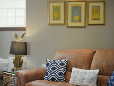 A living room with a brown leather sofa with patterned cushions and three framed artworks above at Budle Granary in Bamburgh
