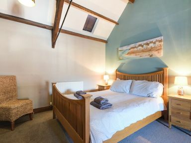 A bedroom with a wooden bed framed by two wooden nightstands with lamps and a wicker chair at Budle Granary in Bamburgh