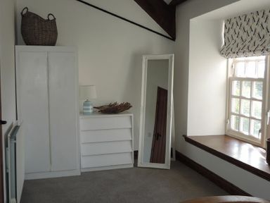 A corner of a room with a white wardrobe a white chest of drawers a standing mirror and a window with a patterned blind at Budle Granary in Bamburgh