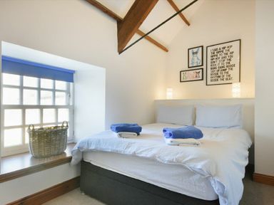 A bedroom with a double bed white bedding and blue towels near a window with a basket and framed pictures on the wall at Budle Granary in Bamburgh