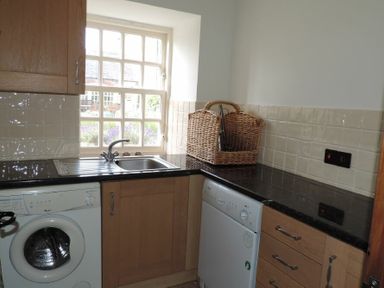 A kitchen area with a washing machine a sink with a window above and a dishwasher next to a wicker basket on black countertops at Budle Granary in Bamburgh