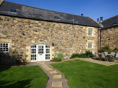 A stone building with white doors and windows and a garden path leading to the door at Budle Granary in Bamburgh
