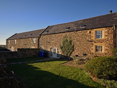 An exterior view of a stone building with a tiled roof and multiple windows a patio area with table and chairs and a grassy garden at Budle Granary in Bamburgh