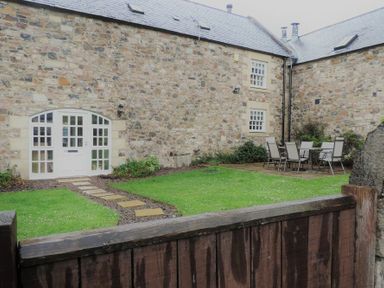 A stone courtyard with a wooden gate a grass lawn a stone pathway glass paneled door and outdoor dining furniture at Budle Granary in Bamburgh