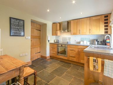 A kitchen with wooden cabinets a wooden door a wooden table and chair and stone tile flooring at Cheshill Cottage in Alnmouth