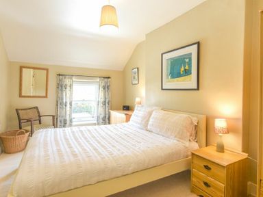 A bedroom with a double bed bedside tables a chair a basket a window with curtains and framed artwork at Cheshill Cottage in Alnmouth