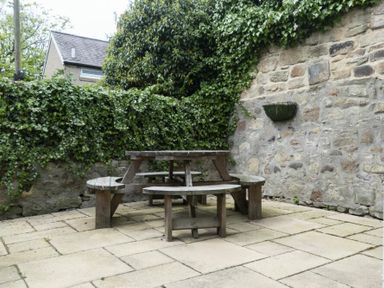 An outdoor patio with a round wooden picnic table surrounded by ivy-covered stone walls at Cheshill Cottage in Alnmouth