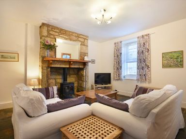 A living room with two white sofas a stone fireplace a television and a window with curtains at Crier Cottage in Alnwick