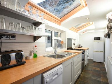 A narrow kitchen with a wooden countertop, sink, dishwasher, stovetop, refrigerator, and glasses on a shelf at Crier Cottage in Alnwick