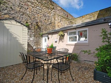An outdoor patio area with a metal table and chairs on a gravel surface next to stone walls at Crier Cottage in Alnwick