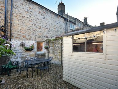 An outdoor patio area with gravel ground a metal table and chairs a green bench hanging flower pots and a cream shed against stone walls at Crier Cottage in Alnwick