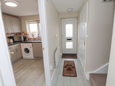 A hallway with a white door and two brown mats next to a kitchen with cabinets a washing machine and a window at Curlew Cottage in Seahouses