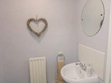 A bathroom with a wall-mounted round mirror a white sink with a bottle of soap a towel ring with a beige towel a heart-shaped wicker wall decoration and a white radiator at Curlew Cottage in Seahouses