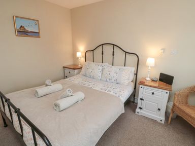 A bedroom with a double bed with floral bedding and rolled towels on top with bedside tables and lamps and a wicker chair at Curlew Cottage in Seahouses