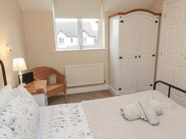 A bedroom with a bed with rolled towels on it a wicker chair a nightstand with a lamp a window and a white wardrobe at Curlew Cottage in Seahouses
