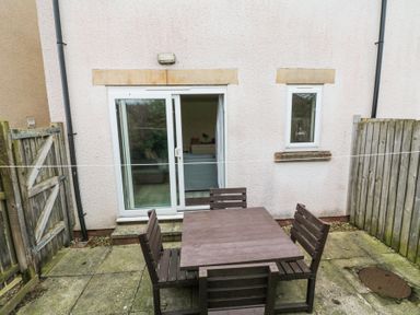 A small patio with a wooden table and four chairs in front of a sliding glass door at Curlew Cottage in Seahouses